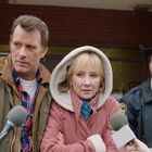 Zwei Menschen stehen vor Mikrofonen; ein Mann umarmt eine Frau in einer Kapuze. Ein Sheriff steht dahinter.