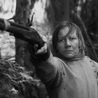 Eine entschlossene Person in altertümlicher Kleidung zielt mit einem Gewehr im Wald, schwarz-weiß gehalten.