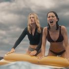 Zwei Frauen auf einem gelben Luftmatratze im Wasser, eine schreit, der Himmel ist bewölkt.