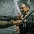 Zwei Männer kämpfen heftig im Regen. Einer hält einen Stock, der andere verteidigt sich mit der Hand.