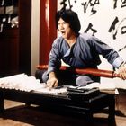 Chinesische Schriftzeichen im Hintergrund. Eine Person in grauer Kleidung sitzt, hält einen roten Gegenstand und zeigt Emotionen.