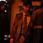 Ein Mann in einem Anzug steht im Regen, umgeben von Personen in Regenmänteln. Die Szene wirkt angespannt und dunkel.