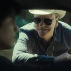 Ein Mann mit Cowboy-Hut und Sonnenbrille beugt sich lächelnd zum Fahrerfenster eines Autos.