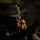 Ein Mann in einem Cowboyhut zielt mit einer Pistole. Die Szene ist dunkel und dramatisch beleuchtet.