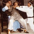 Drei Männer in Kampfposition. Einer sitzt erschrocken auf einem Stuhl, während die anderen beiden in Aktion um ihn herum sind.