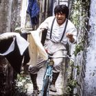 Ein Mann fährt erschrocken mit einem Fahrrad durch eine enge Gasse mit hängender Wäsche und Pflanzen an den Wänden.