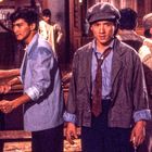 Rotes Banner mit asiatischen Schriftzeichen, Männer in Vintage-Kleidung in einem Raum, teilweise in intensiven Gesprächen.