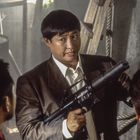 Ich kenne den Text nicht. Ein Mann im Anzug hält eine Waffe, umgeben von weiteren Personen in einer Werkstatt.