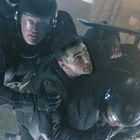 Drei Männer in futuristischer Kleidung; einer wird von zwei anderen festgehalten. Sie tragen Helme und Schutzanzüge.