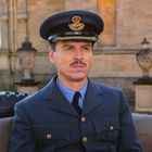 Ein Mann in einer blauen Uniform mit Flügelabzeichen und Mütze sitzt auf einem Sofa vor einem historischen Gebäude.