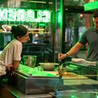 Neon-Schriftzug "CLAREMON" im Fenster. Eine Frau lehnt an einer Theke, während ein Mann Pizza zubereitet.
