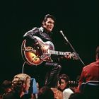 Ein Mann in schwarzem Lederanzug sitzt auf einem Hocker, spielt Gitarre und lächelt; Menschen sitzen um ihn herum.