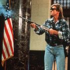 Ein Mann in Sonnenbrille und kariertem Hemd hält ein Gewehr. Neben ihm weht eine US-Flagge in einem Gebäude.