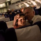 Ein Mann und eine Frau sitzen im Inneren eines Flugzeugs. Der Mann hält die Frau beruhigend. Beide wirken besorgt.