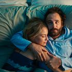 Ein Mann und eine Frau liegen in einem Bett, sie kuscheln sich aneinander. Der Mann trägt ein blaues Pyjamahemd.
