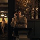 Ein Mann mit gestreiftem Pullover telefoniert, steht neben einem dekorativen Holzmuster. Im Hintergrund sind Leute in einem Restaurant.