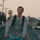 Ein junger Mann fährt lächelnd auf einem Fahrrad eine Straße entlang, umgeben von Autos und Palmen im Hintergrund.