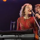 Zwei Personen singen gemeinsam in Mikrofone. Die Frau trägt ein rotes, glitzerndes Kleid, der Mann ein dunkles Hemd.