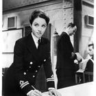 Eine Frau in Marineuniform beugt sich über einen Tisch, während zwei Männer im Hintergrund stehen und sitzen.