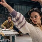 Eine Frau in einem weißen Pullover tanzt fokussiert in einem Atelier; im Hintergrund ein Mann an einem Tisch.