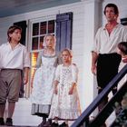 Mehrere Personen in historischer Kleidung stehen auf einer Veranda. Ein Mann und fünf Kinder, alle schauen ernst.