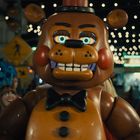Großer, orange-brauner Animatronik-Bär mit schwarzer Fliege, umgeben von leuchtenden Lichtern und schaulustigen Menschen.