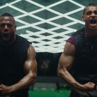 Zwei muskulöse Männer schreien mit angespannten Muskeln, einer trägt ein Shirt mit Nike-Logo. Helles Licht fällt von oben.