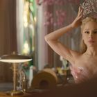 Eine Frau in schulterfreiem Kleid setzt sich eine Krone auf, vor einem Spiegel in einem stilvollen Raum.
