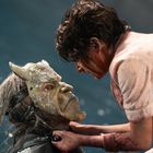 Eine Person mit schmutzigem Gesicht hält eine maskierte Person fest. Die Maske ähnelt einem dämonischen Gesicht mit Hörnern.