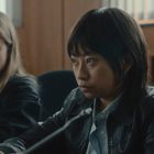 Zwei Personen sitzen an einem Tisch, eine mit blonden Haaren und die andere mit dunklem Pony, konzentriert und ernst schauend.