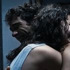 Eine Umarmung zwischen zwei Personen mit lockigem Haar in einem dunklen Raum. Regal mit Büchern im Hintergrund.