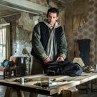 Es sind keine lesbaren Texte zu erkennen. Ein Mann in einer heruntergekommenen Wohnung packt einen Rucksack auf einem Tisch.