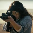 Eine Person, konzentriert mit einem Gewehr zielend, trägt Schutzbrille; im Hintergrund eine verschwommene Landschaft.