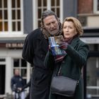 Zwei Personen stehen draußen, schauen in einen Amsterdam-Reiseführer. Sie sind warm gekleidet, Gebäude im Hintergrund.