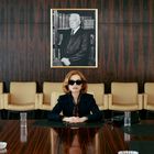 Ein Porträt an der Wand. Eine Frau mit Sonnenbrille sitzt an einem Tisch, umgeben von leeren Stühlen in einem Konferenzraum.