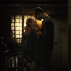 Text auf einer Tür: "MOORE'S". Ein Mann und eine Frau stehen sich nahe in einem schmalen, dunklen Ziegelgang.