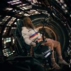 Ein bärtiger Mann in einem Cockpit, umgeben von leuchtenden Instrumenten. Er trägt eine weiße Jacke mit bunten Streifen.