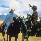 Eine Frau in blauem Umhang und ein Mann auf Pferden, umgeben von Menschen in traditioneller Kleidung in einer Landschaft.