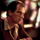 Ein Mann in einem Anzug sitzt nachdenklich an einer Bar, umgeben von unscharfem, farbigem Licht.