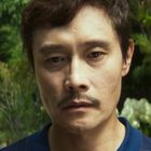 Ein Mann mit ernstem Blick steht im Freien, umgeben von Bäumen und Grünpflanzen. Er trägt einen blauen Hemdkragen.