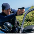 Ein Mann in dunkler Kleidung zielt mit einer Pistole über die Motorhaube eines Autos. Hintergrund zeigt Büsche und Meer.