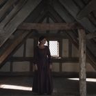 Keine erkennbaren Texte. Eine Frau in einem Holzdachboden, Licht fällt durch ein kleines Fenster.