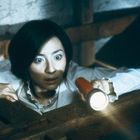 Eine Frau mit besorgtem Ausdruck schaut mit einer Taschenlampe in einen dunklen Raum.