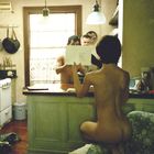 Text: "John Currin." Eine nackte Person sitzt auf einem Küchenstuhl, schaut Richtung Küchentresen, wo zwei weitere Personen stehen.