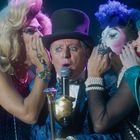 Eine Gruppe von Menschen flüstert einem Mann mit Zylinder und Mikrofon Geheimnisse ins Ohr.