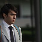 Ein junger Mann im grauen Anzug und schwarzer Krawatte trägt einen blauen Rucksack und schaut nachdenklich zur Seite.