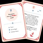 Zwei Karten: Erste Karte fragt "Was passiert, wenn man Puhee macht?" mit drei Optionen: Feiern, Sich aufregen, Schlitten fahren. Zweite Karte erklärt "Puhee" als Aufregung. Verspieltes Layout mit nautischen Symbolen und rotem Rand.