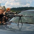 Es gibt keinen gut lesbaren Text zu erwähnen. Zwei Personen zielen mit einem Gewehr über die Motorhaube eines Autos.