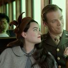 Ein Paar sitzt in einem historischen Zug, sie lächeln einander an. Er trägt eine Militäruniform, sie einen braunen Hut.