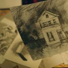 Eine Hand hält eine Zeichnung eines Hauses, umgeben von weiteren Zeichnungen auf einem Tisch.